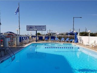 A bright and sunny outdoor pool at a hotel, with blue umbrellas, sun loungers, and a clear blue sky